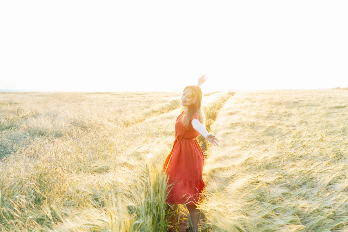 Gabriele Harder, Eine glückliche junge Frau mit erhobenen Armen entspannt sich bei Sonnenuntergang im Weizenfeld